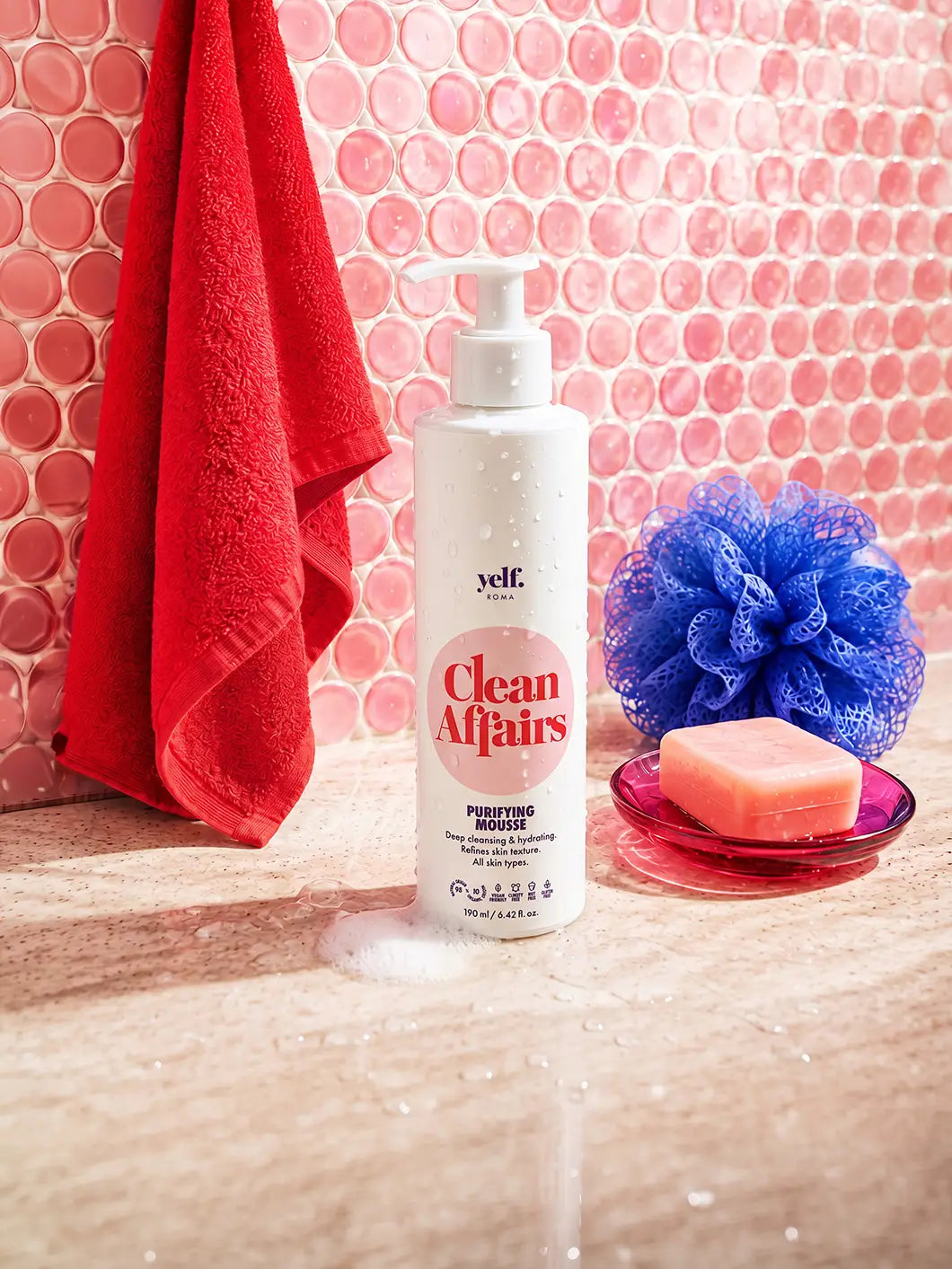 Yelf Clean Affairs Purifying Mousse in a vibrant bathroom scene with pink circular glass mosaic tiles, red towel, blue shower sponge and pink soap dish on a wet stone surface.
