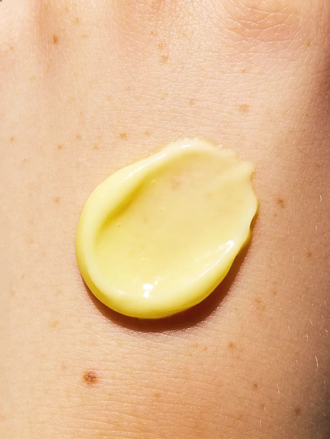 Close-up of a thin lemon-yellow vitamin C serum layer on very light skin with subtle freckles, showing a lightweight, silky skincare texture