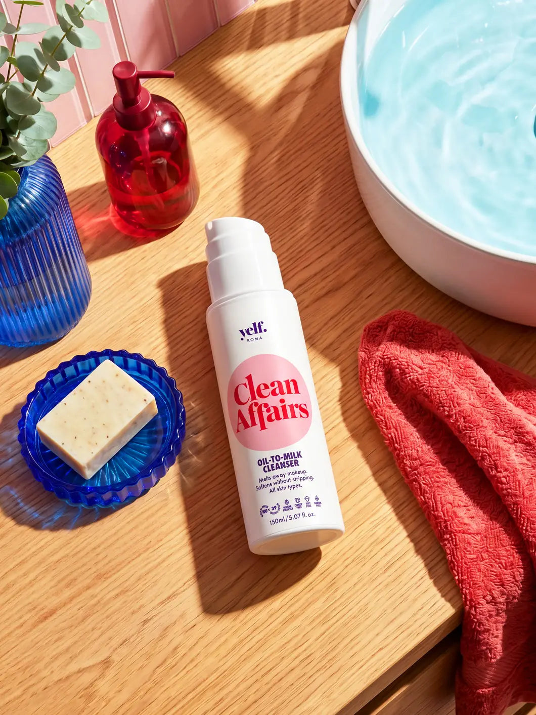 Top-view lifestyle image of Yelf Clean Affairs Oil-to-Milk Cleanser on a wooden bathroom vanity beside a light blue sink and coral towel in bright natural sunlight.