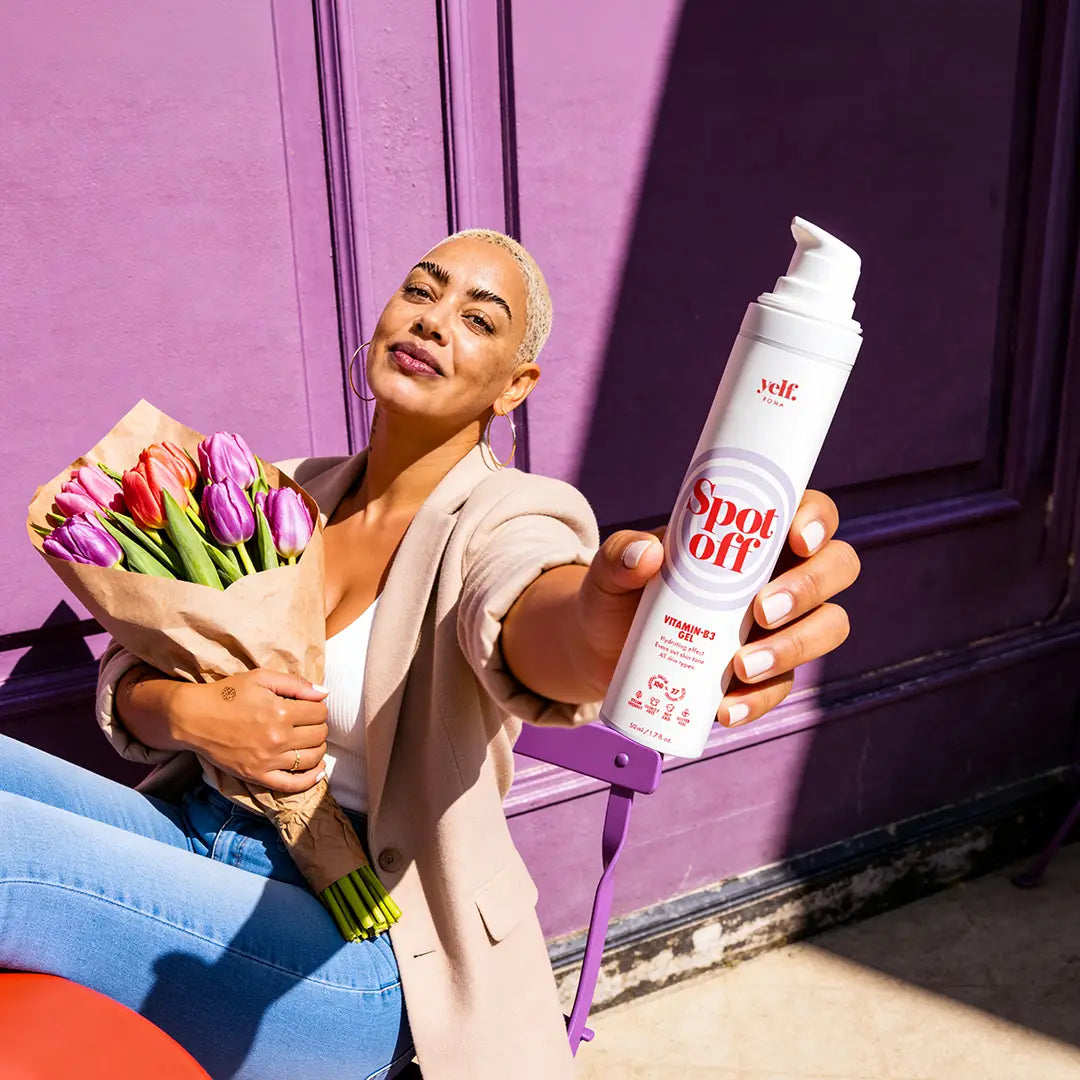 Model holding Yelf Spot Off Vitamin-B3 Gel while sitting outdoors in bright sunlight with colorful flowers, vibrant purple backdrop and playful summer city aesthetic.