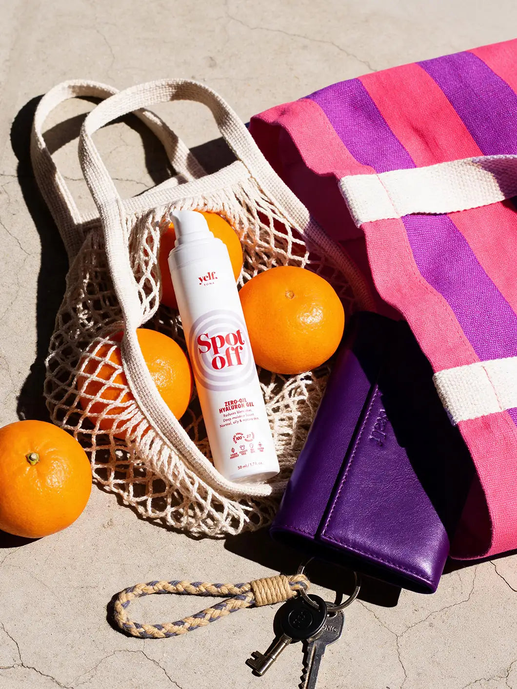 Yelf Spot Off Zero-Oil Hyaluron Gel with oranges in a mesh bag, striped tote bag and wallet on concrete in bright sunlight