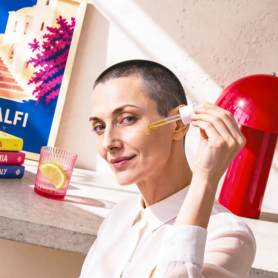 Woman applying facial oil with a dropper in a bright editorial home interior with warm sunlight and colorful modern design accents.