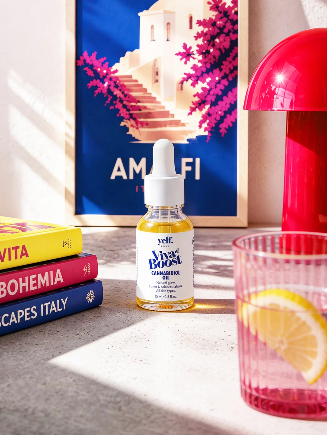 Yelf Viva el Boost Cannabidiol Oil in a bright Mediterranean lifestyle scene with Amalfi poster, pink lamp, lemon water glass and colorful books on a concrete surface