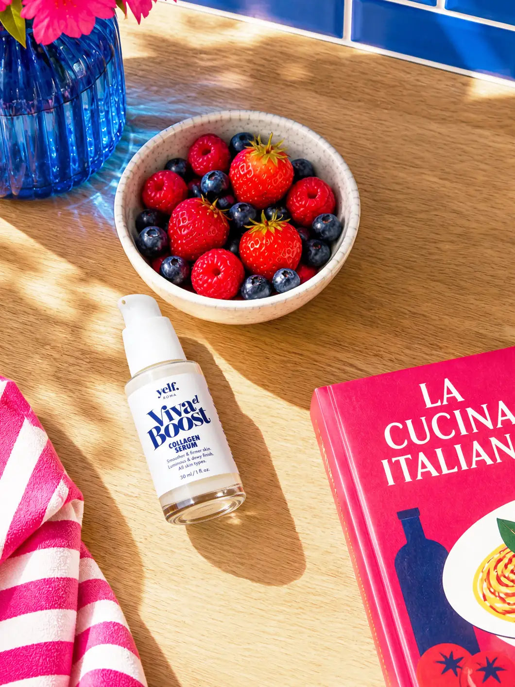 Yelf Viva el Boost Collagen Serum lying on a sunlit wooden kitchen surface with fresh berries, lemon water, pink striped towel and blue glass vase in a bright, high-contrast lifestyle scene